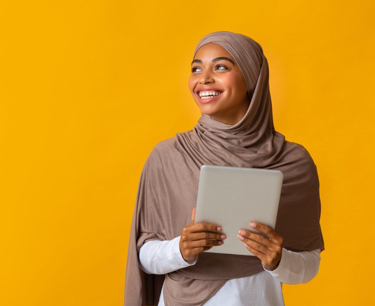Images shows a smiling black muslim girl in hijab holding digital tablet