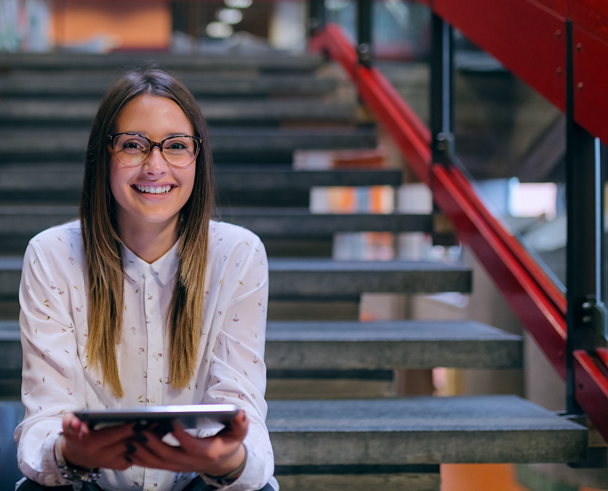 Image show girl with glasses smiling and sitting on a step in a contemporary setting using her tablet device.