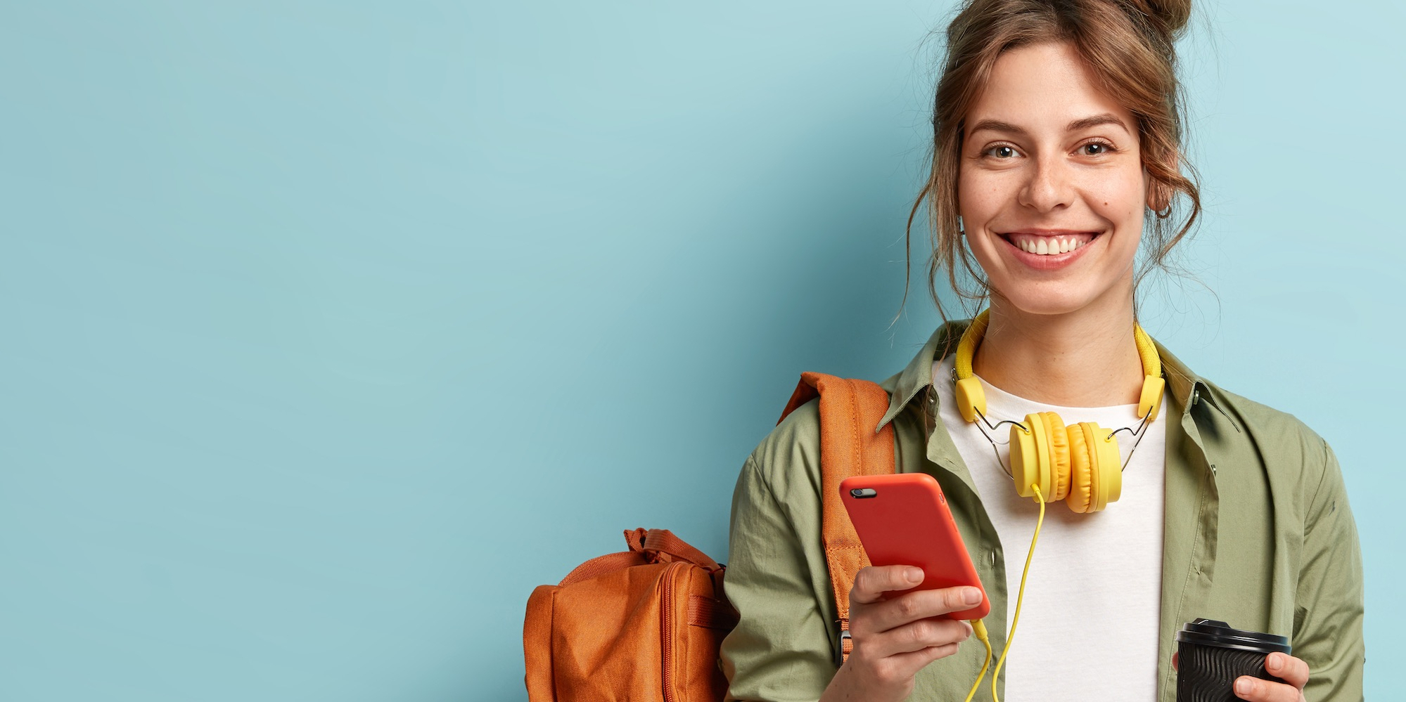 Image shows a smiling student with a rucksack over her shoulder, a coffee in one hand and her mobile device in the other.